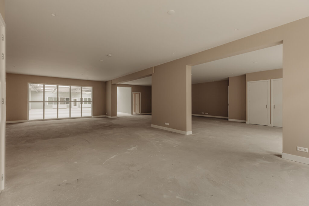 Ruime, lege binnenruimte aan de Henri Polaklaan met beige muren en grote ramen naar een atrium.